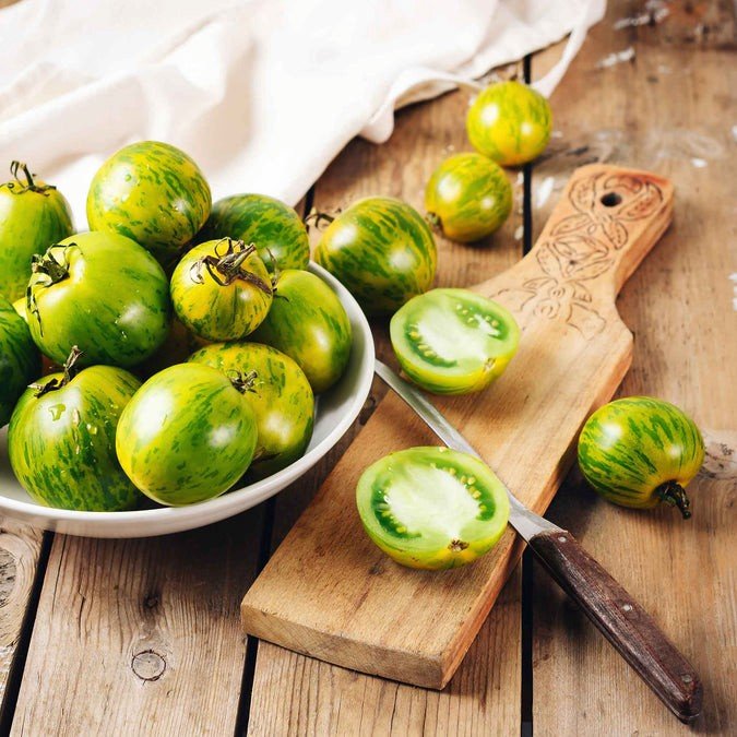 Fresh green heirloom tomatoes in a bowl with a sliced tomato on a wooden cutting board and a knife, showcasing their vibrant color and unique striped pattern.